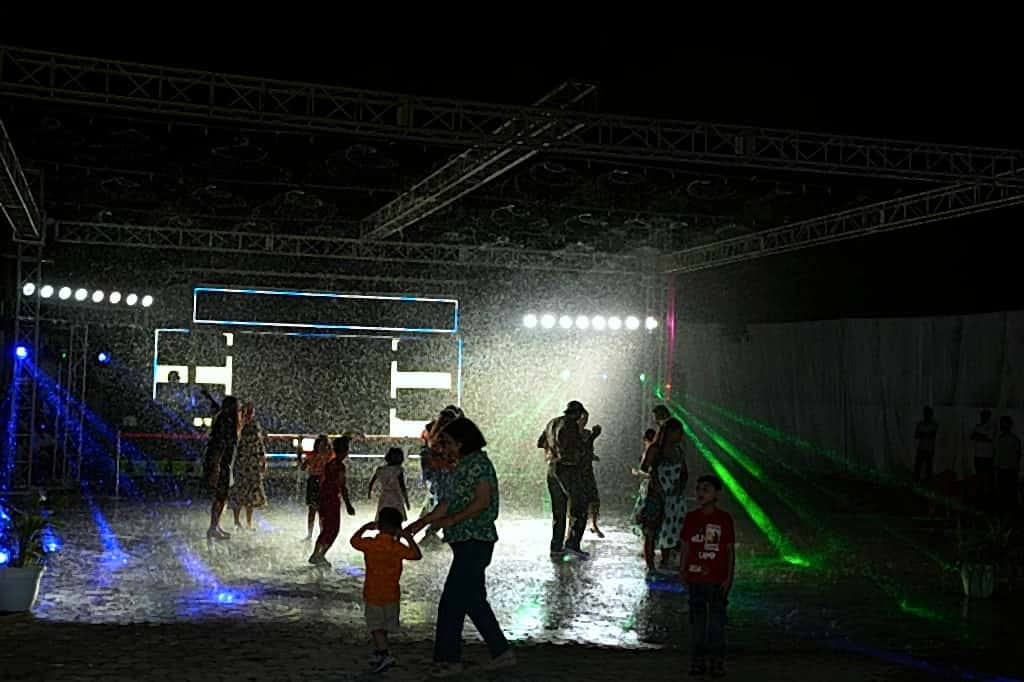 Guests and children dancing under illuminated water spray on an outdoor rain-dance floor