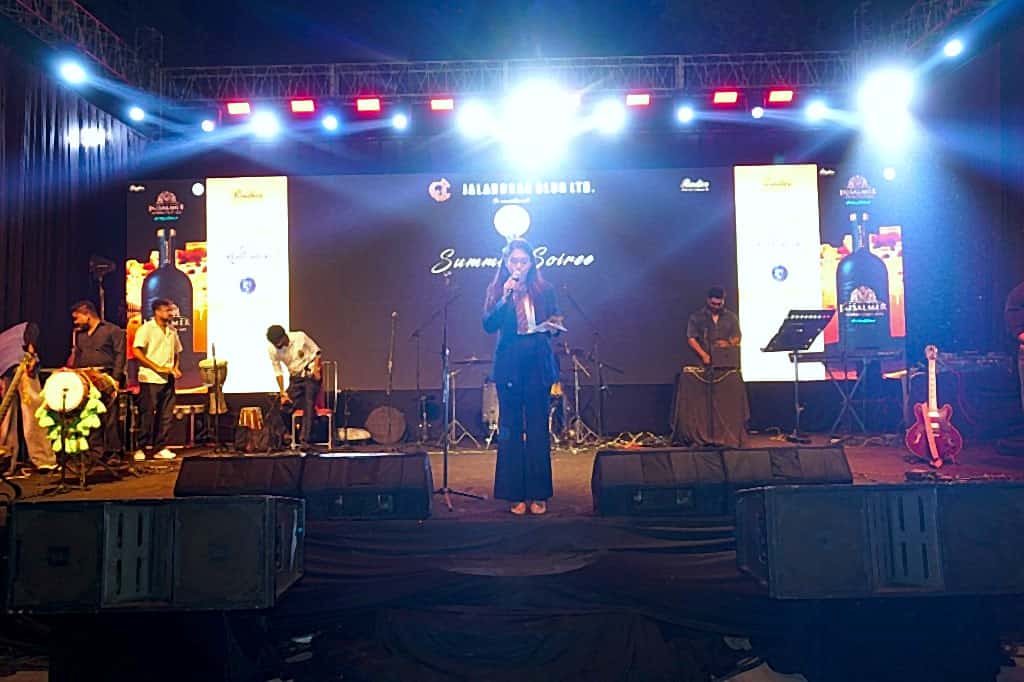 Host addressing guests on a lit outdoor stage with band, DJ, and LED backdrop
