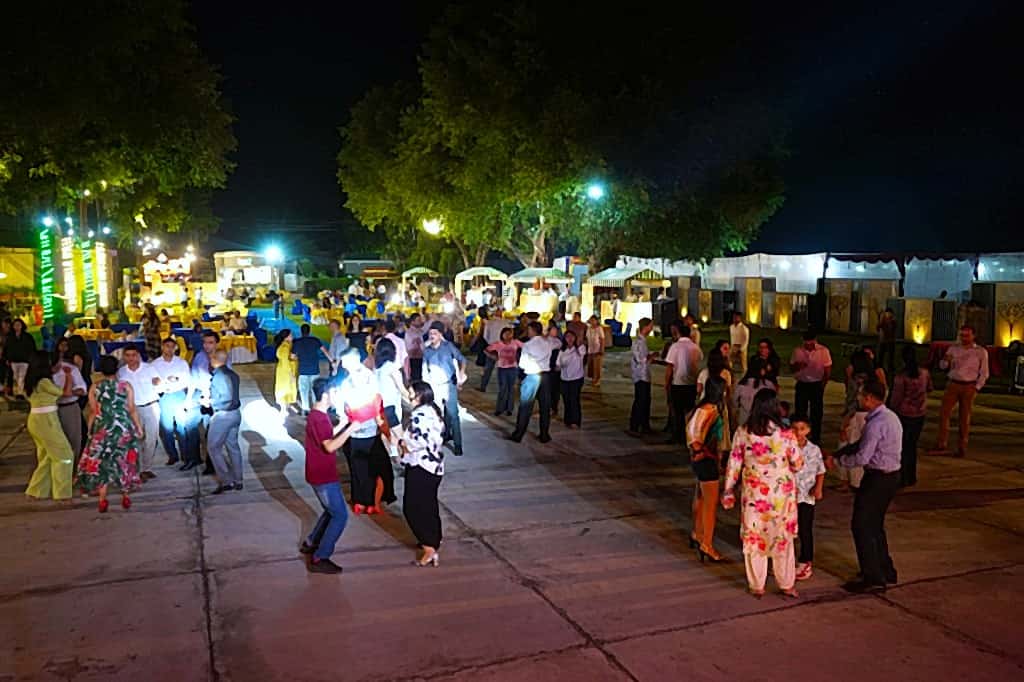 Large outdoor social gathering dancing under trees and decorative lighting