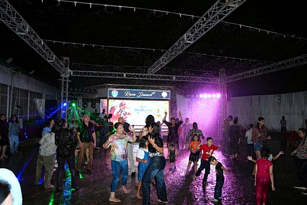 Crowd dancing in purple-lit artificial rain under outdoor truss and LED stage